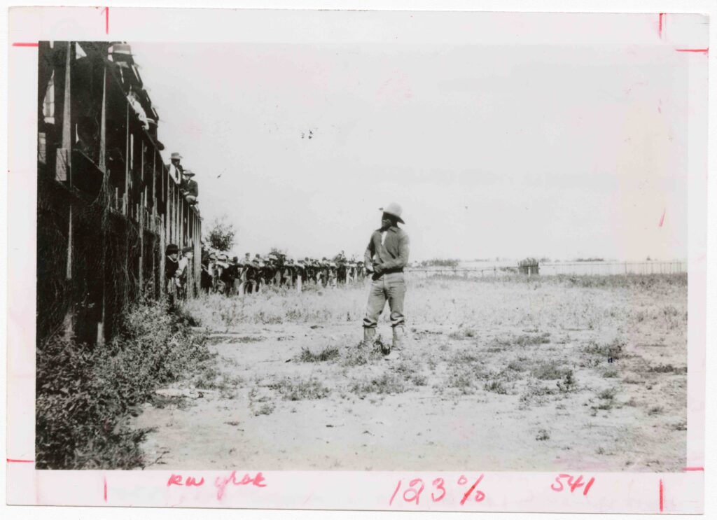 Bill Pickett stands in front a wooden platform. A crowd of people can be seen in the distance. 